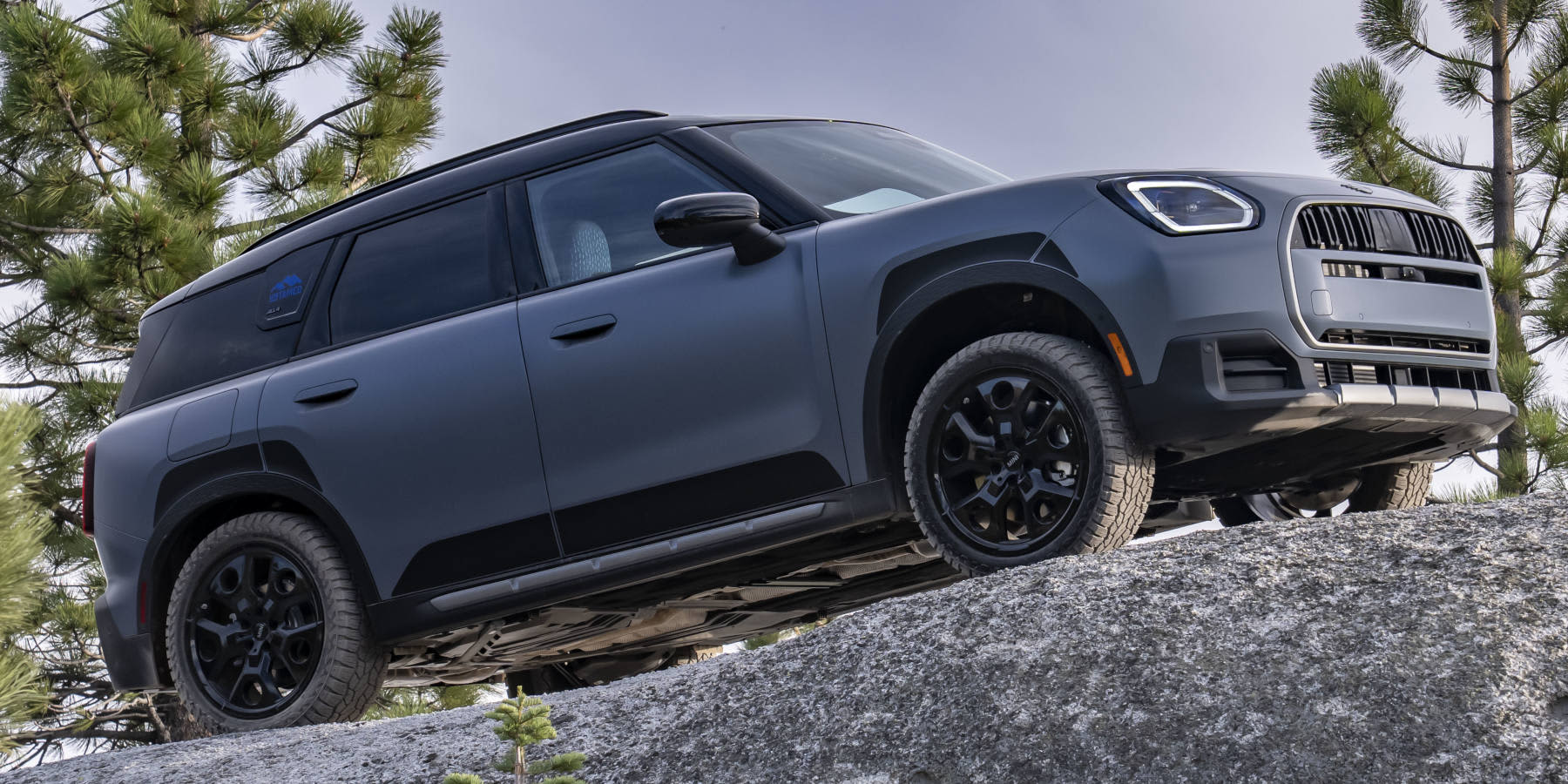 Ein mattgrauer MINI Countryman ALL4 steht auf einem felsigen Untergrund vor Nadelbäumen. Prägend sind der markante Achteck-Kühlergrill, prägnante Scheinwerfer und die robuste Offroad-Silhouettte.   
