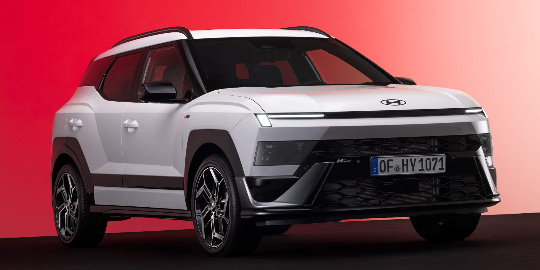 Ein weißer Hyundai Bayon N Line steht vor rotem Hintergrund. Auffällig sind die durchgehende LED-Lichtleiste, der sportliche Kühlergrill und markante Radhäuser.   