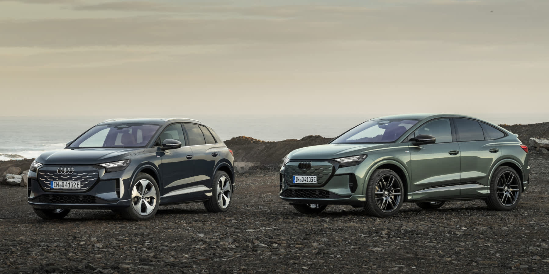 Das Bild zeigt zwei moderne Elektro-SUVs von Audi, die nebeneinander auf einem steinigen, unebenen Untergrund an einer Küste stehen. Es handelt sich um Modelle der Audi Q4 e-tron Baureihe.