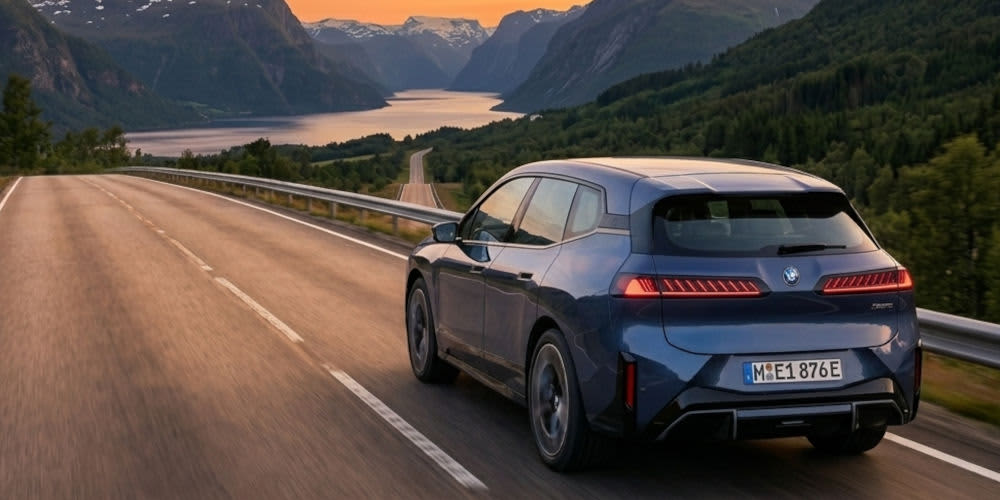Ein blauer BMW iX3 fährt auf einer Straße durch eine norwegische Fjordlandschaft im Abendlicht. Das Elektro-SUV ist von hinten zu sehen und hat ein Münchener Kennzeichen (M EE 876E).