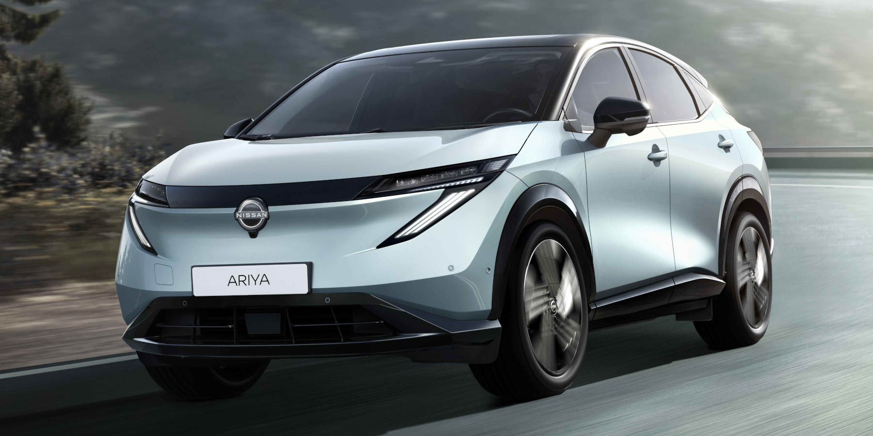 Ein hellblauer Nissan Ariya in Fahrt auf einer Straße. Das Elektro-SUV besticht durch schmale LED-Scheinwerfer, ein schwarzes Dach und das markante „Ariya“-Kennzeichen vor bergiger Kulisse.