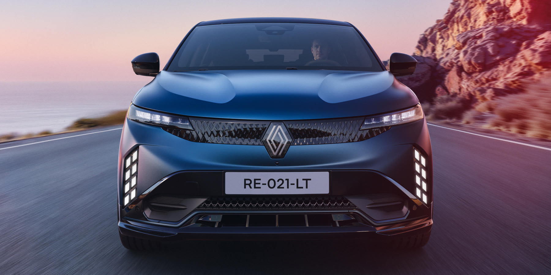 Ein blauer Renault Megane E-Tech elektrisch fährt frontal auf einer Straße an einer Küste. Das Auto hat auffällige vertikale Tagfahrlichter, schmale Scheinwerfer und das Kennzeichen RE-021-LT.