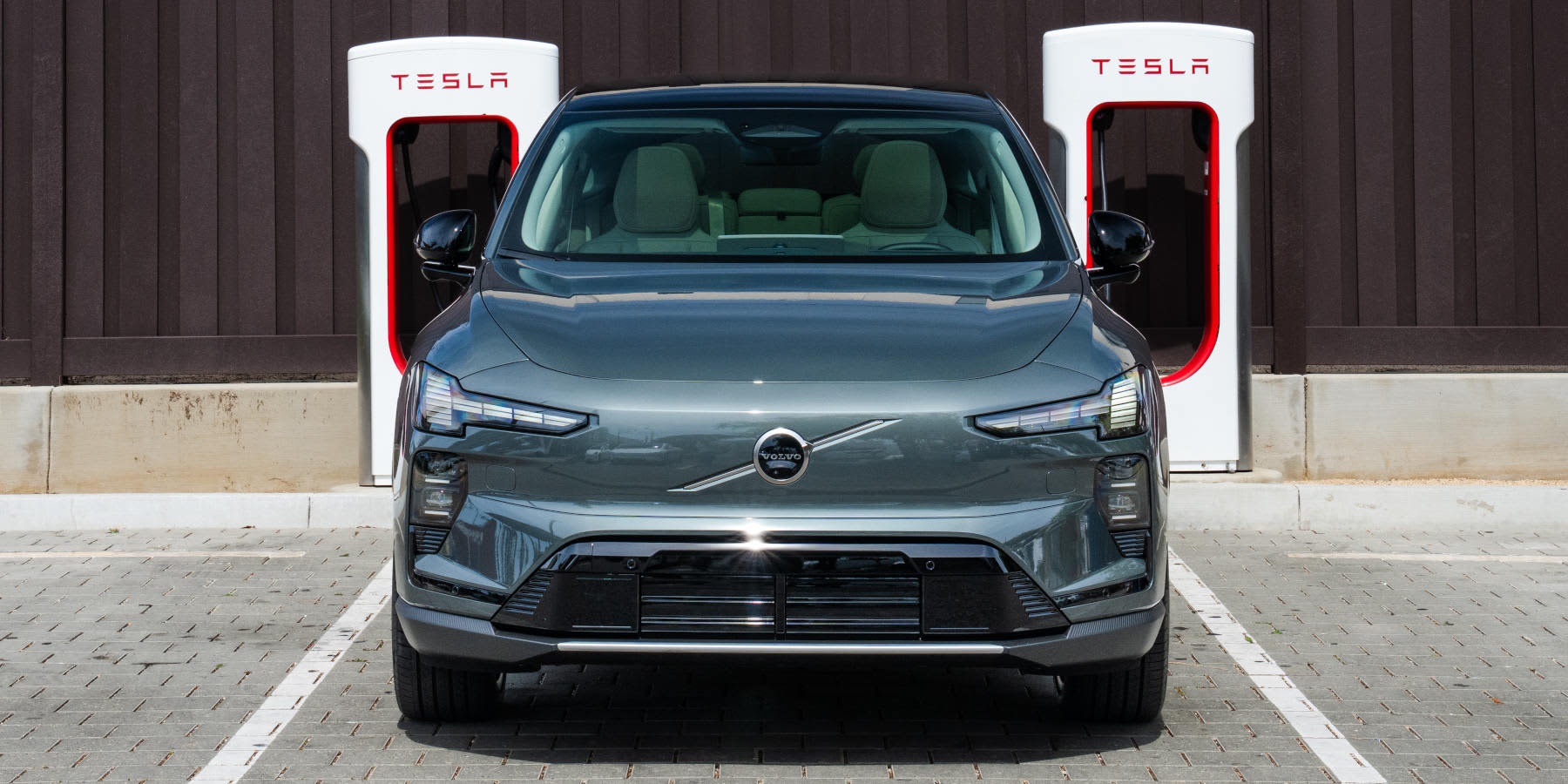 Ein dunkelgrauer Volvo steht frontal auf einem Parkplatz vor zwei weißen Tesla Supercharger-Ladesäulen.