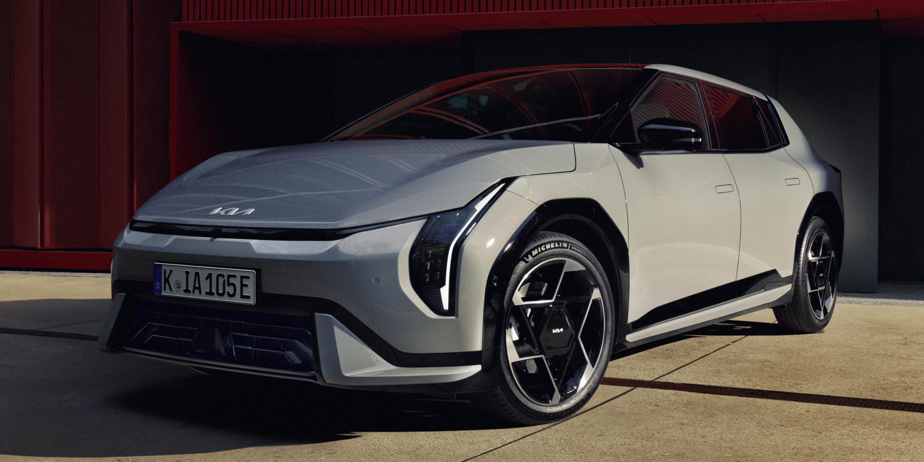 Schrägansicht eines grauen Kia EV4 vor rotem Gebäude. Das Elektrofahrzeug zeigt vertikale LED-Scheinwerfer, auffällig geometrische Felgen und das deutsche Kennzeichen K IA 105E.