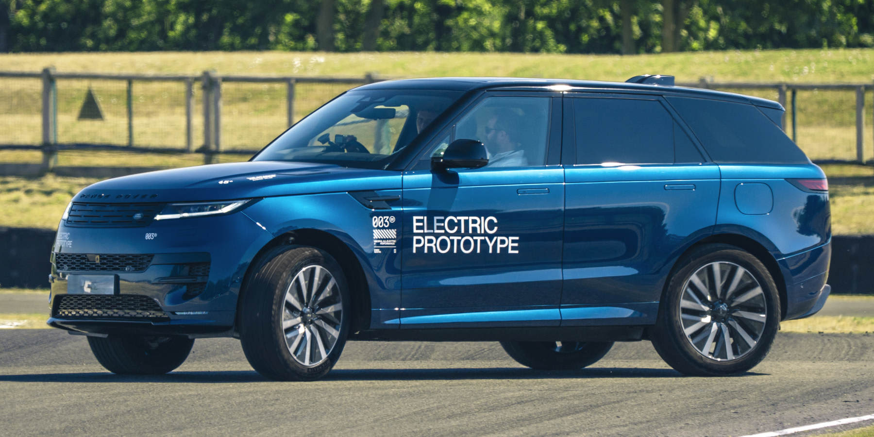 Ein blauer Range Rover Prototyp mit der Aufschrift „ELECTRIC PROTOTYPE“ fährt auf einer Teststrecke vor einer grün bepflanzten Kulisse. (KI-generiert)