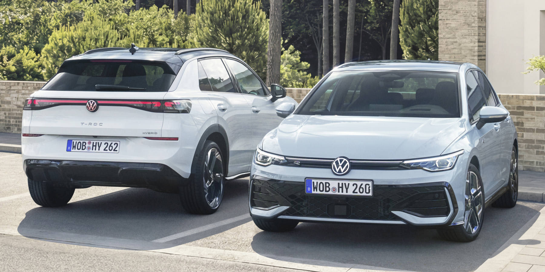 Zwei hellgraue VW-Modelle parken nebeneinander: Ein T-Roc Hybrid von hinten und ein Golf Hybrid von vorne, vor einer Kulisse aus Bäumen und Mauern.