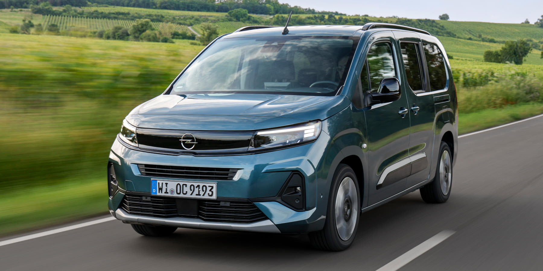 Ein blaugrauer Opel Combo fährt dynamisch auf einer Landstraße entlang. Im Hintergrund erstrecken sich grüne Felder und Hügel unter leicht bewölktem Himmel.