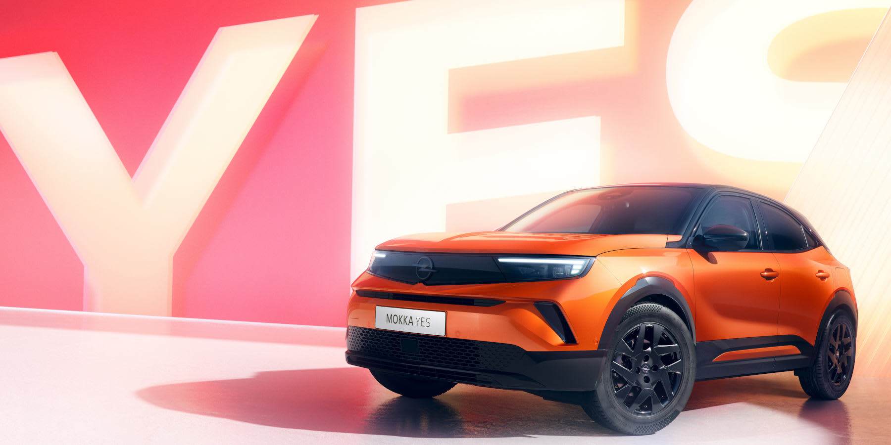 Ein leuchtend orangefarbener Opel Mokka YES mit schwarzem Dach steht vor einer rosafarbenen Wand mit großem, leuchtendem „YES“-Schriftzug. (KI-generiert)