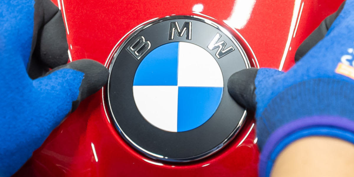 Hände in blauen Schutzhandschuhen montieren das runde BMW-Emblem präzise auf einer glänzend roten Oberfläche. (KI-generiert)