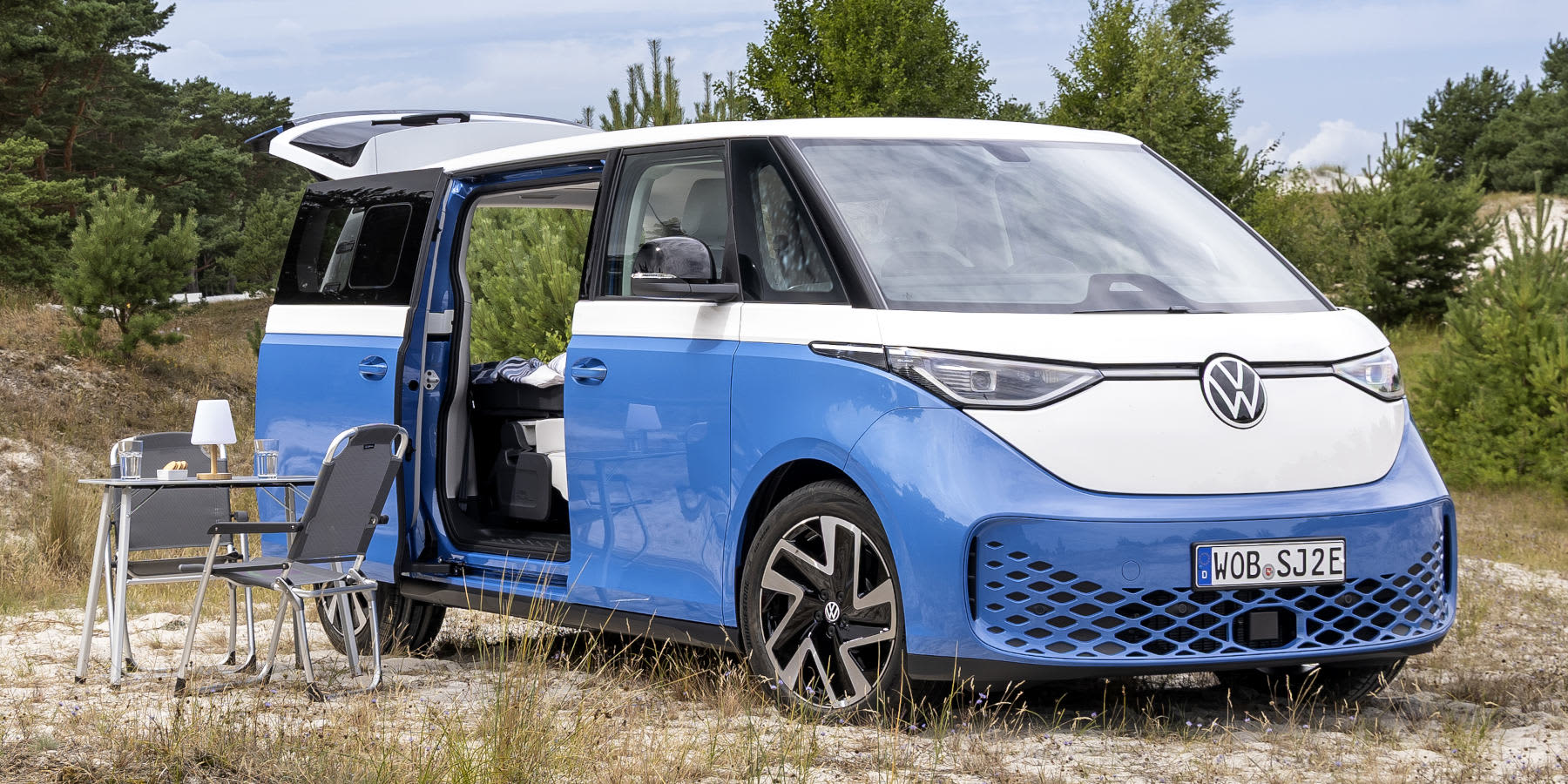 Ein blau-weißer VW ID. Buzz Camper Van steht in der Natur mit geöffneter Schiebetür, aufgestellter Heckklappe und Campingmöbeln daneben. (KI-generiert)