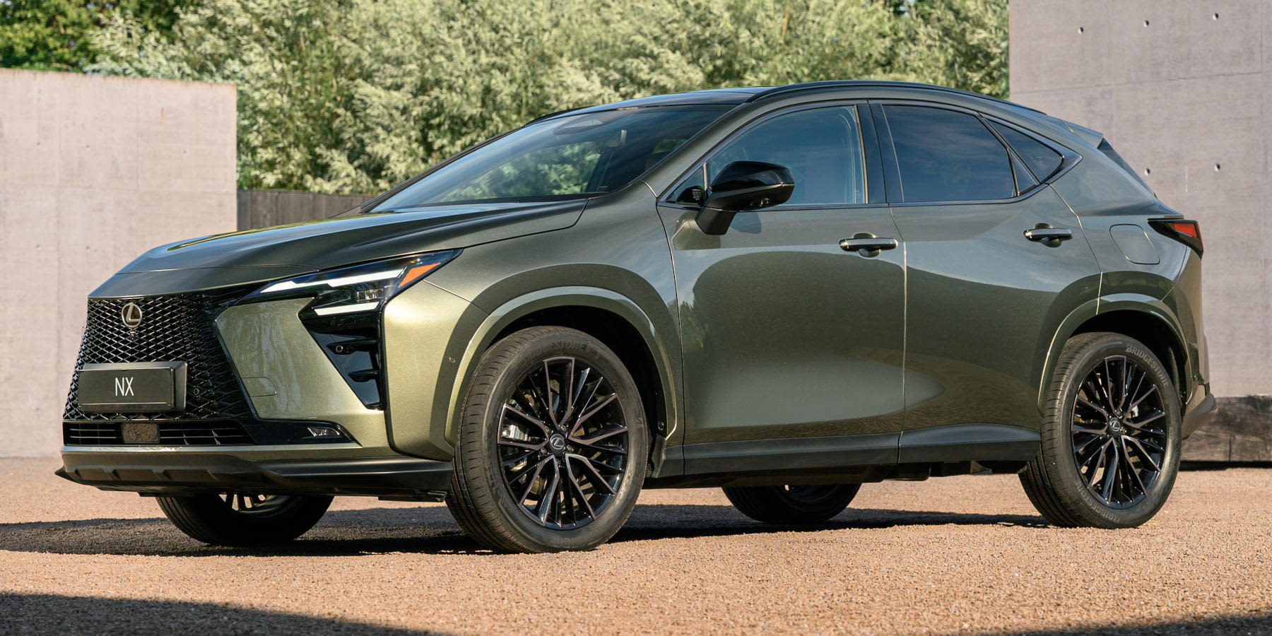 Ein olivgrüner Lexus NX SUV steht auf einem Kiesuntergrund vor einer Betonwand. Das Fahrzeug hat einen auffälligen Wabengrill, scharfe LED-Scheinwerfer und schwarze Felgen. (KI-generiert)