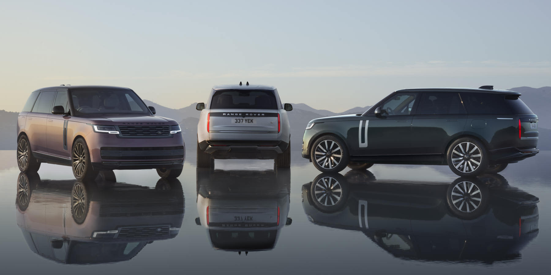 Drei Range Rover Electric SUVs stehen nebeneinander auf einer spiegelnden Fläche vor einer Bergkulisse und zeigen Front-, Heck- und Seitenansicht.