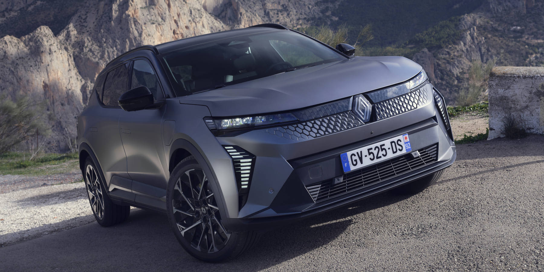 Ein mattgrauer Renault Scénic E-Tech Electric steht auf einer Straße vor einer Felslandschaft. Auffällig ist der strukturierte Grill. (KI-generiert)