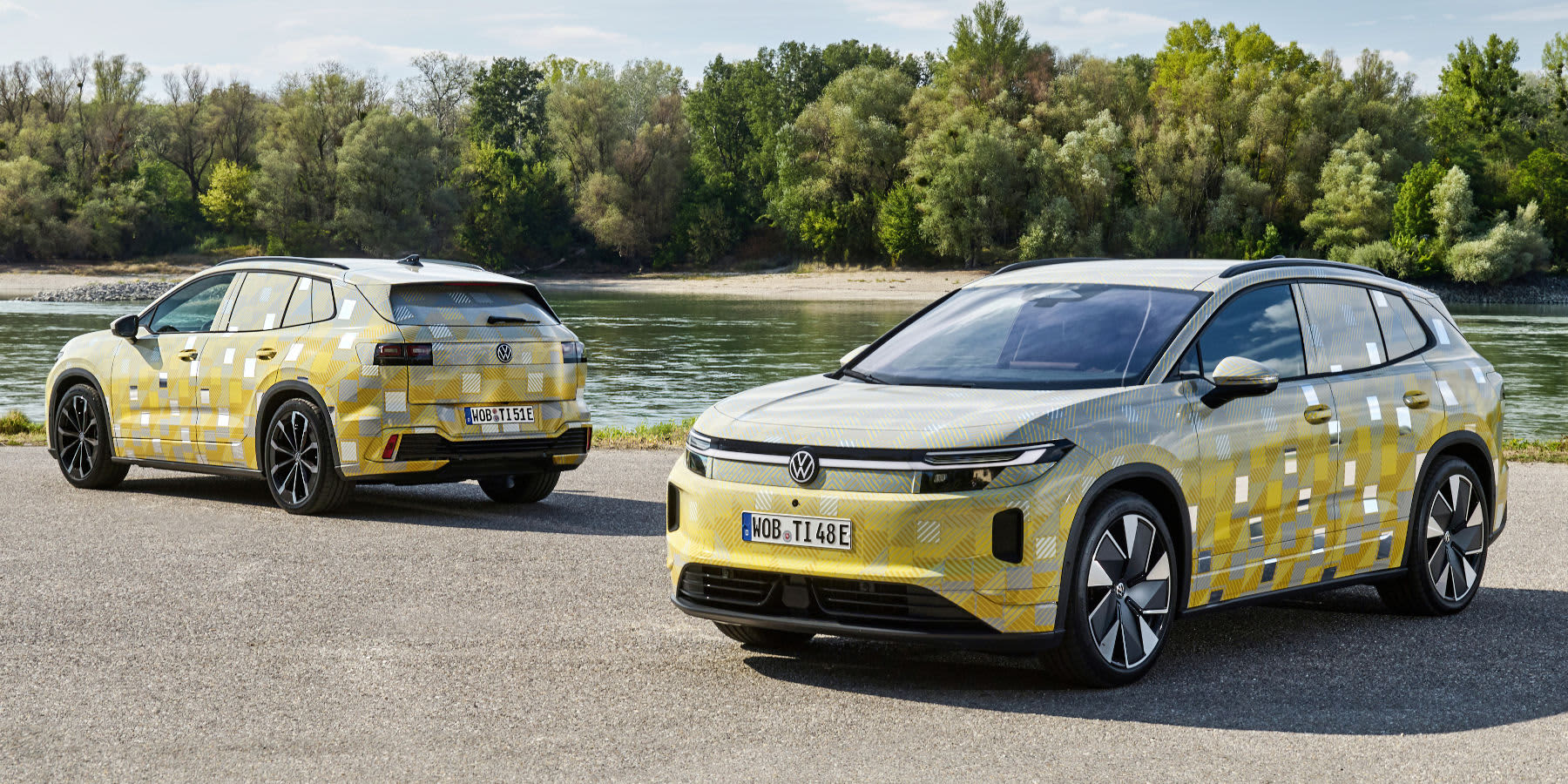 Zwei getarnte VW ID. Tiguan-Erlkönige stehen an einem Flussufer. Prägend sind das gelb-graue Tarnmuster, das durchgehende Lichtband und die SUV-Proportionen. (KI-generiert)