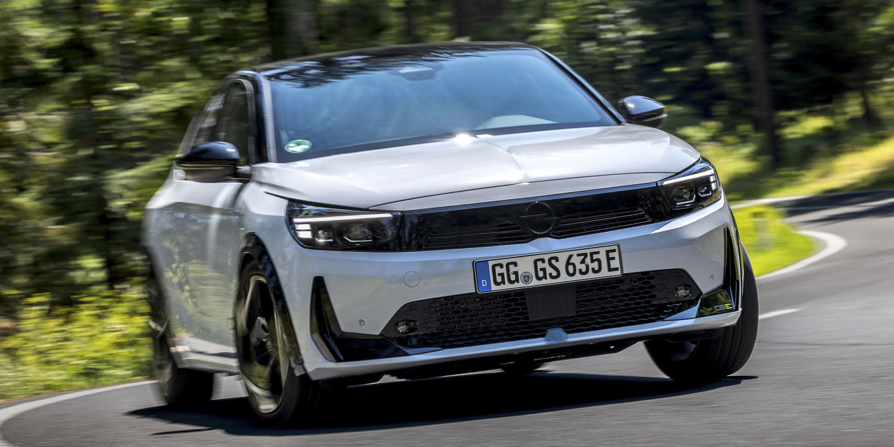 Ein silbergrauer Opel Corsa GSE fährt dynamisch auf einer schattigen Waldstraße. Markant sind das schwarze Visor-Grillgesicht und das schwarze Dach. (KI-generiert)
