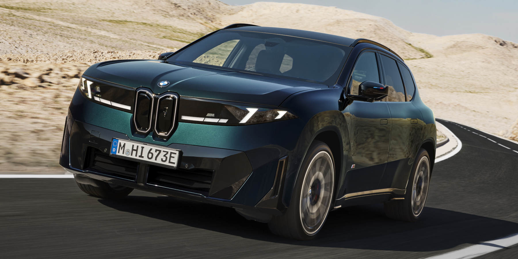 Ein dunkler BMW iX3 M60 xDrive fährt dynamisch auf einer Bergstraße. Prägend sind die beleuchtete Niere, die neue Lichtsignatur und die athletische SUV-Silhouette.   