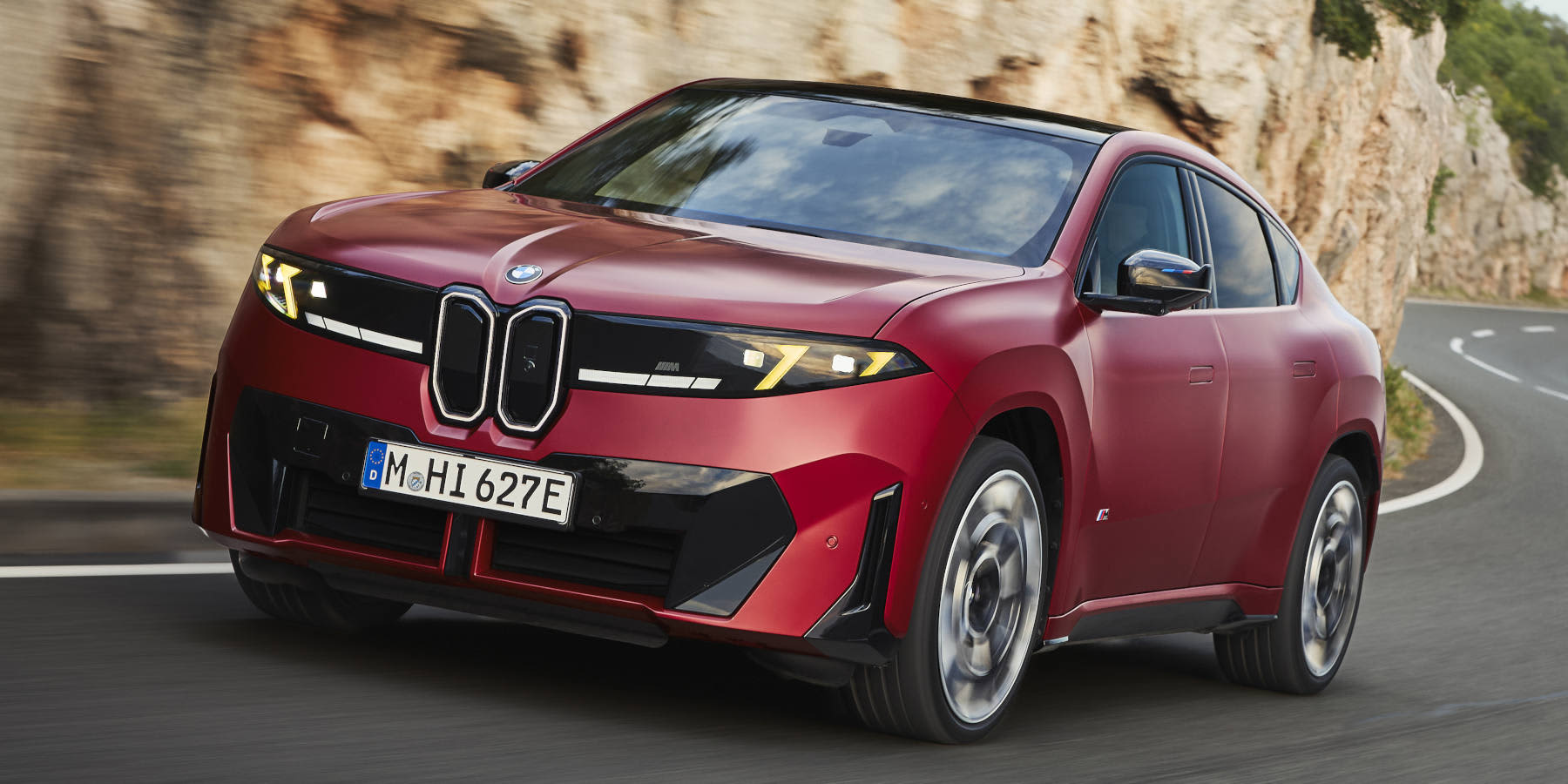 Ein roter BMW iX4 M60 xDrive fährt dynamisch an einer Felswand entlang. Prägend sind die beleuchtete Niere, die neue Lichtsignatur und die SUV-Coupe-Form.   