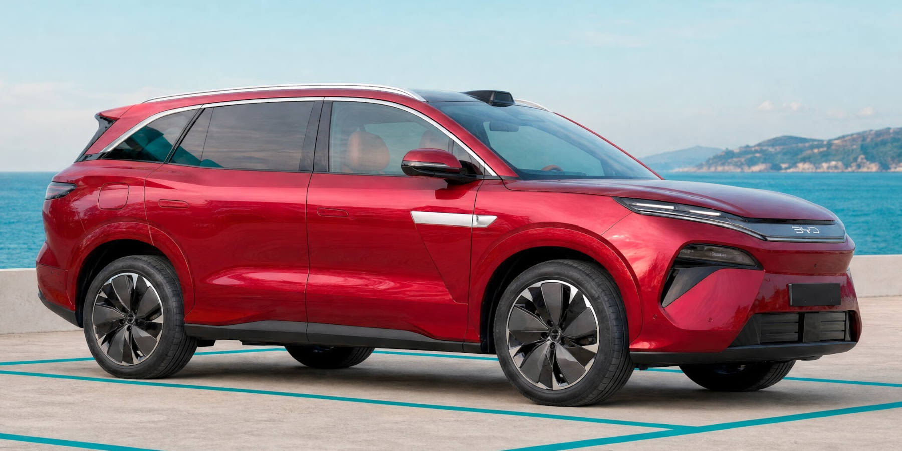Ein roter BYD Atto 8 steht vor einer Küstenkulisse am Meer. Das SUV fällt durch seine gestreckte Silhouette, schmale LED-Lichter und große Alufelgen auf.   