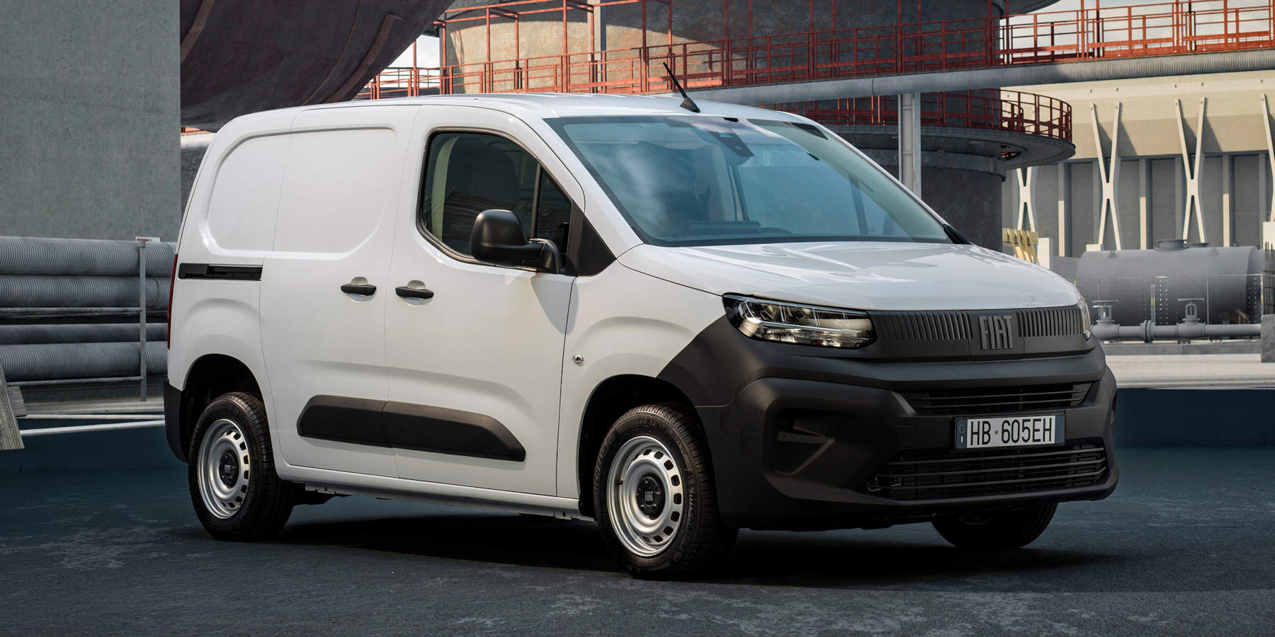 Ein weißer Fiat Doblo steht vor einer Industrieanlage. Prägend sind der robuste, unlackierte Stoßfänger, das schmale Scheinwerferband und der FIAT-Schriftzug.   