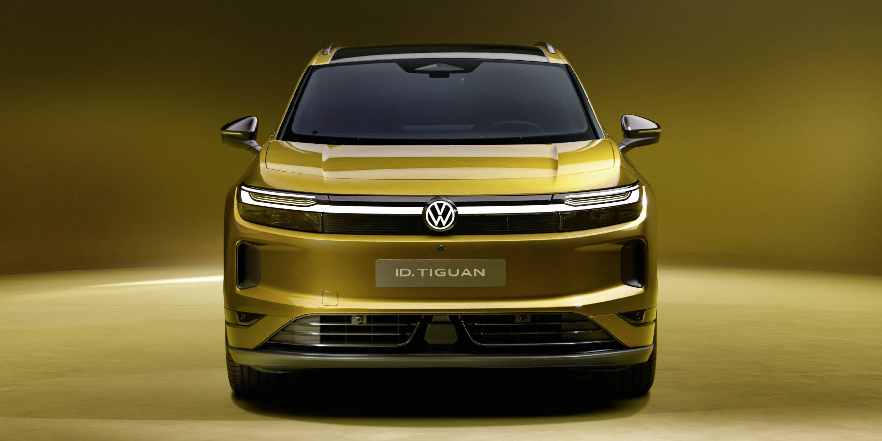 Ein goldener Volkswagen ID. Tiguan wird frontal im Studio präsentiert. Prägend sind das durchgehende LED-Lichtband, das beleuchtete VW-Logo und die breite Frontansicht.   